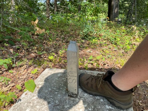 A person’s foot in a hiking boot next to a metal boundary marker on a forested trail. Green foliage surrounds the area.