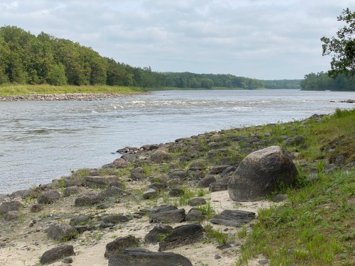 A calm river flows beside a rocky shore, surrounded by lush green trees under a cloudy sky.