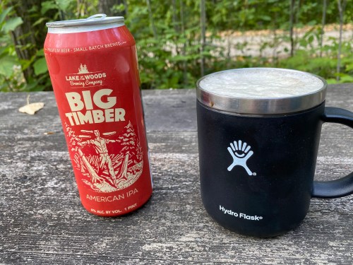 A can of Big Timber American IPA next to a black Hydro Flask mug on a wooden surface in a natural setting.
