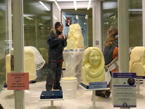 A sculptor carves a butter statue while another person sits nearby, both in a display case at a fair.