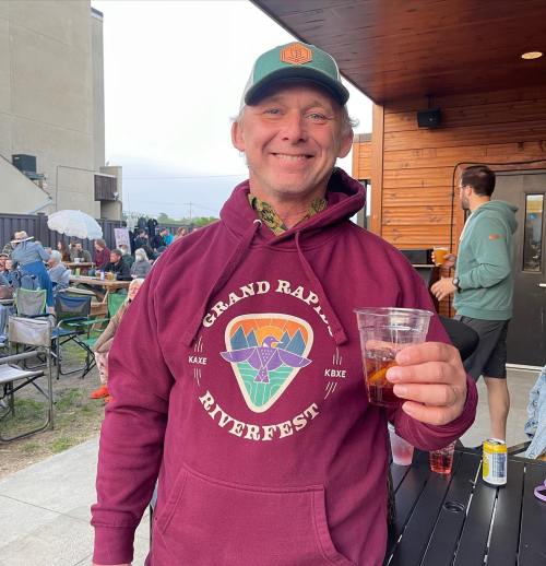 A smiling man in a maroon hoodie holds a drink at an outdoor event with people and tents in the background.