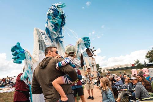 A man holds a child as they watch a large, colorful puppet at an outdoor event with an audience in the background.