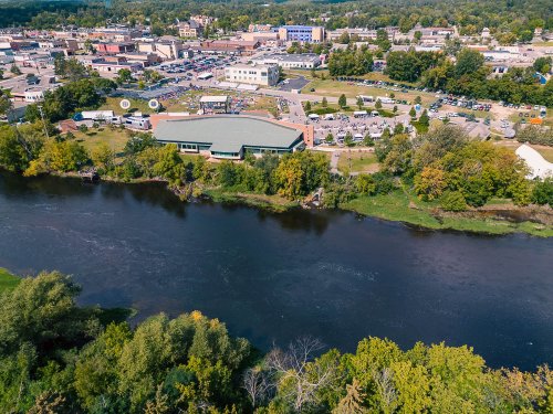 Aerial view of a river alongside a town, featuring green spaces, buildings, and parked cars in a vibrant landscape.