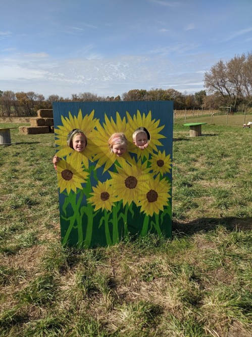 Three children peek through a sunflower-themed cutout in a grassy field on a sunny day.