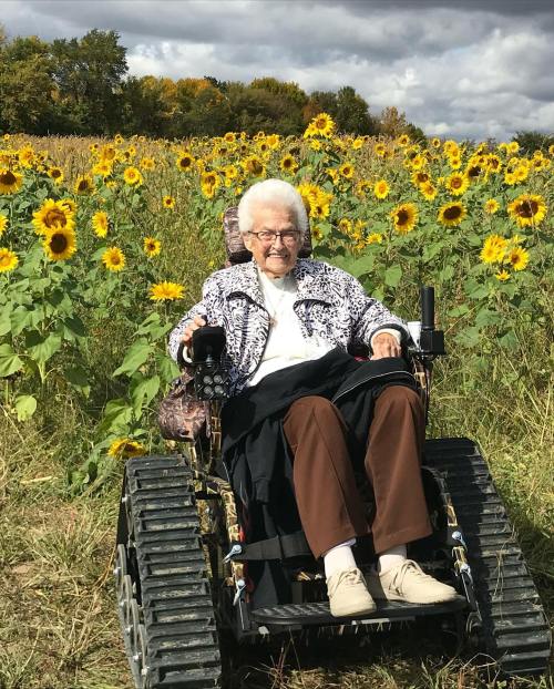 An elderly woman in a wheelchair sits among vibrant sunflowers under a cloudy sky.