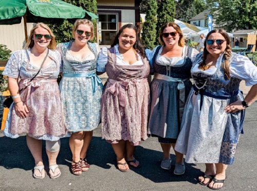 Five women in traditional dresses pose together outdoors, smiling and enjoying a festive atmosphere.
