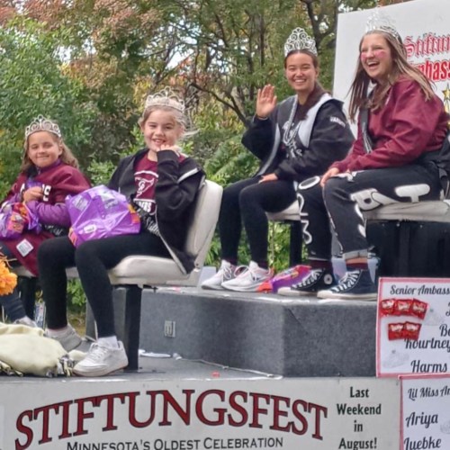 Four girls wearing crowns and jackets wave from a float at a parade, celebrating Stiftungsfest in Minnesota.