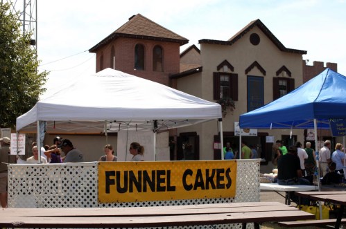 A food stall with a "Funnel Cakes" sign at a lively outdoor event, surrounded by tents and people.