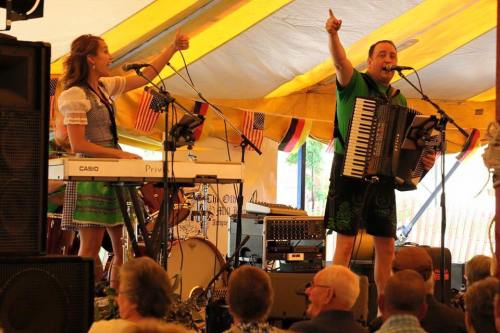 A lively performance under a tent, featuring a woman singing and a man playing accordion, with an audience enjoying the show.