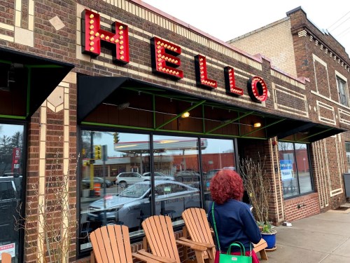 A person with curly red hair stands outside a brick building with a large "HELLO" sign and wooden chairs.