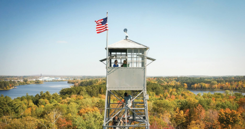 A fire tower with two people at the top, surrounded by colorful autumn trees and a scenic lake view.