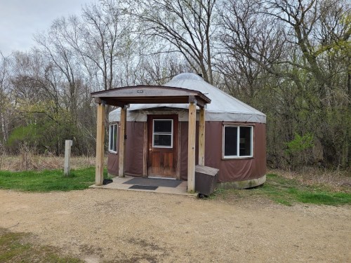 A yurt with a wooden porch, surrounded by trees and grass, set in a natural outdoor environment.