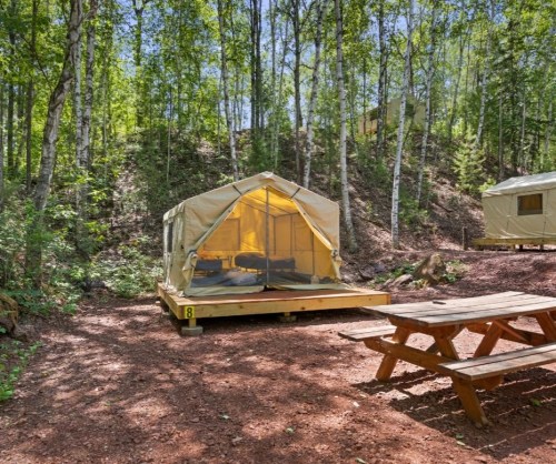 A tent on a wooden platform in a forested area, with a picnic table nearby and trees in the background.