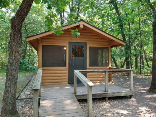 A wooden cabin with a porch, surrounded by trees in a lush green forest.