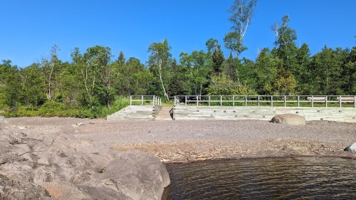 A rocky shoreline with a person walking near wooden steps, surrounded by trees under a clear blue sky.