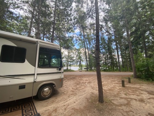 A parked RV in a forested area, with a view of a lake and cloudy sky in the background.