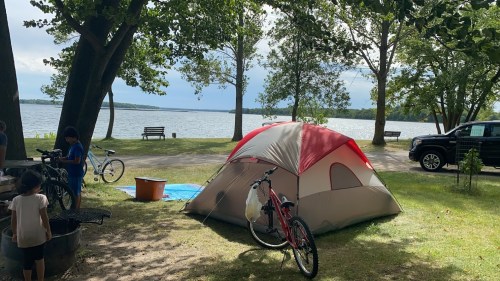 A campsite by a lake with a tent, bicycles, and trees, featuring a picnic area and a parked vehicle in the background.