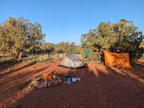 A campsite with a tent, a campfire, cooking supplies, and a tarp set up among trees in a sunny outdoor setting.