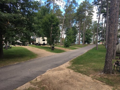 A peaceful campsite with gravel paths, surrounded by tall trees and green grass, leading to a lake in the distance.