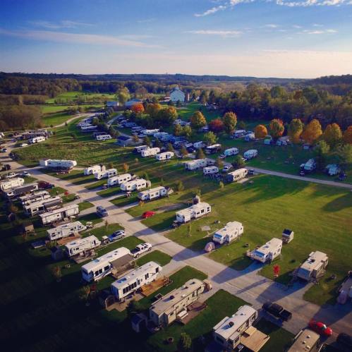 Aerial view of a campground with numerous RVs parked on green grass, surrounded by trees and rolling hills.