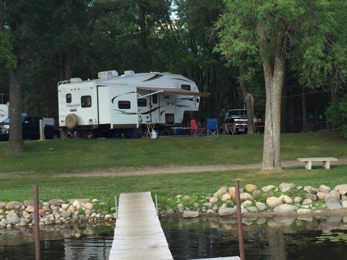 A camper parked by a lake, with chairs set up nearby and a wooden dock leading to the water. Trees surround the area.