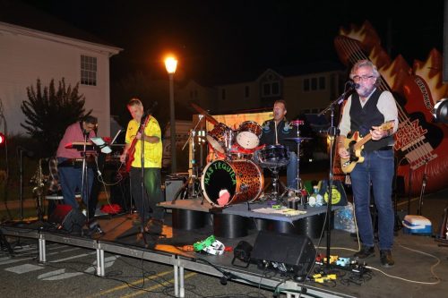 A band performs on stage at night, featuring a guitarist, drummer, and other musicians with instruments.