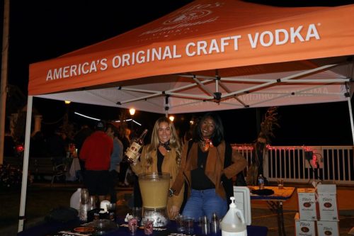 Two women stand behind a table with drinks under an orange tent that reads "America's Original Craft Vodka."