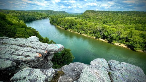 A scenic view of a river winding through lush green hills, seen from a rocky overlook under a partly cloudy sky.
