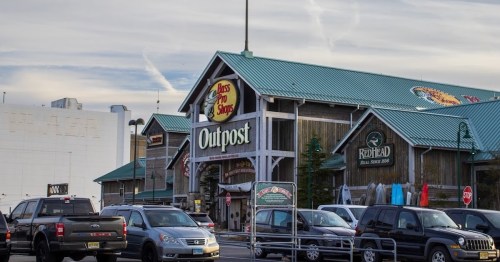 A large outdoor retail store with a green roof, featuring a sign that reads "Bass Pro Shops Outpost" and parked cars.