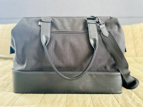 A black duffel bag with leather accents and a shoulder strap, resting on a textured surface.