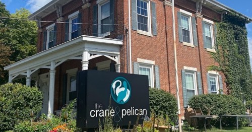A brick building with a porch and a sign reading "the crane & pelican," surrounded by greenery and flowers.