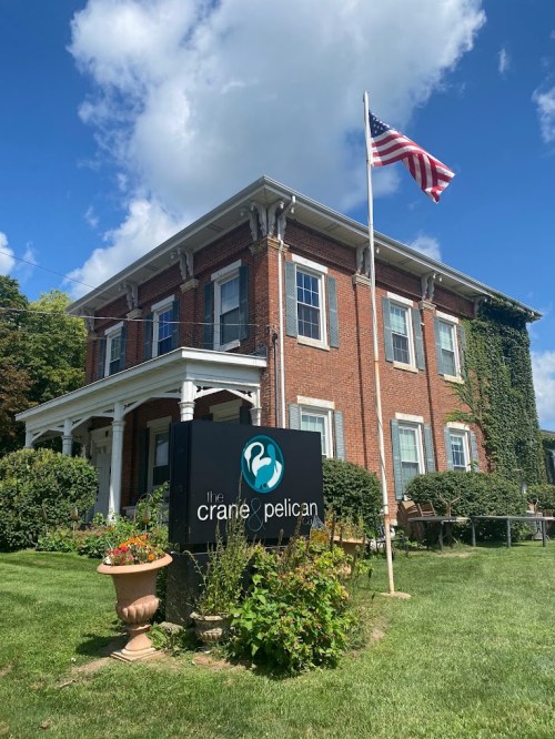 A brick building with a sign reading "the crane & pelican," surrounded by greenery and featuring an American flag.