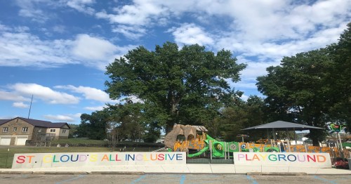 Colorful sign for "St. Cloud's All Inclusive Playground" with playground equipment and blue sky in the background.