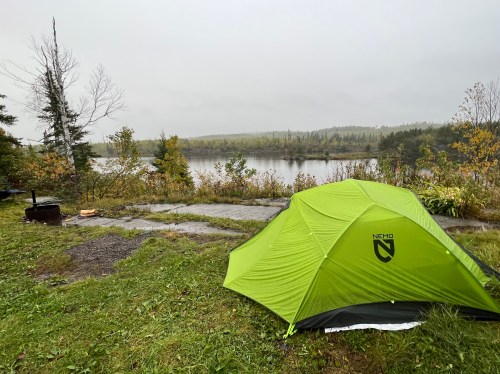 A green tent set up by a calm lake surrounded by trees, with a cloudy sky and wet ground.