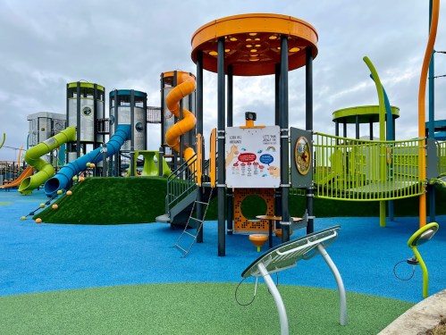 Colorful playground with slides, climbing structures, and interactive panels on a blue and green surface under a cloudy sky.