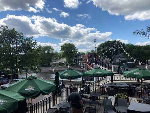 View of a riverside patio with green umbrellas, string lights, and a bridge in the background under a partly cloudy sky.