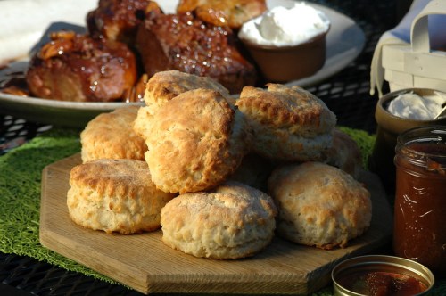A stack of golden biscuits on a wooden platter, with barbecue dishes and condiments in the background.