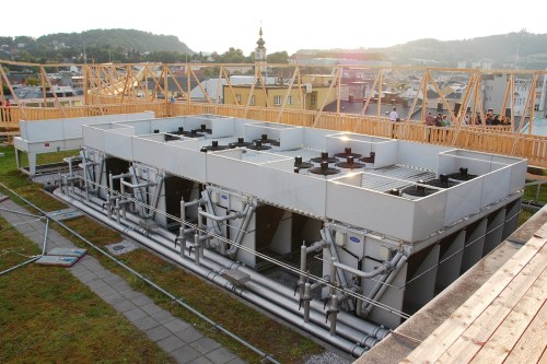 A rooftop installation featuring large cooling units with pipes, surrounded by a wooden structure and cityscape in the background.