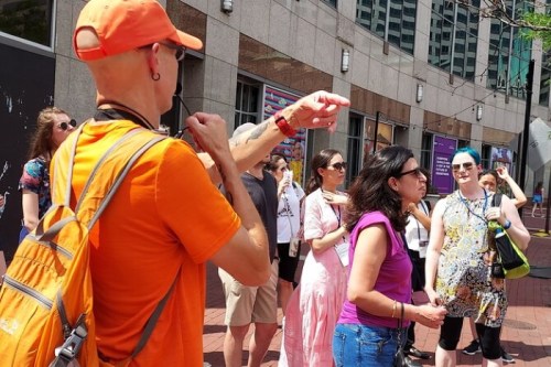 A diverse group of people gathered outdoors, listening to a guide in an orange shirt, with buildings in the background.