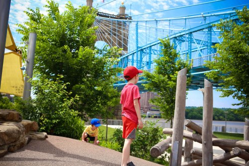 Two children play in a park with a blue bridge in the background and green trees under a bright sky.