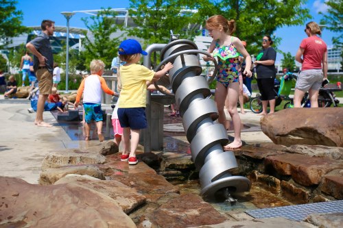 Children play at a water feature in a park, enjoying the sun and splashing water on a warm day.