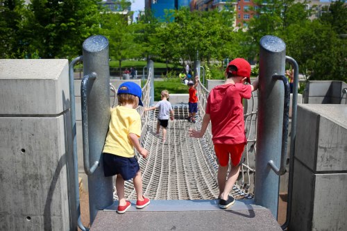 Two children in colorful outfits play on a rope bridge in a park, with other kids visible in the background.