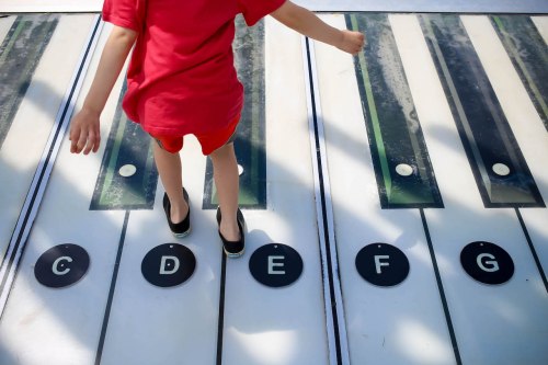 A child in a red shirt plays on a large piano keyboard, stepping on the keys labeled C, D, E, F, and G.