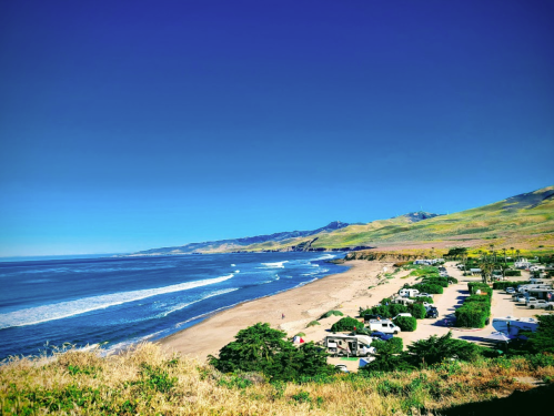 A scenic view of a beach with gentle waves, green hills, and a clear blue sky, featuring nearby camping sites.