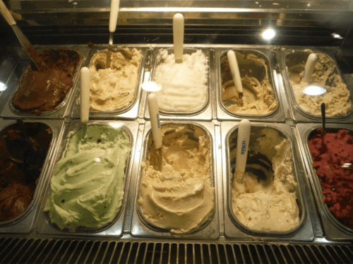 A display of various ice cream flavors in metal tubs, with spoons in each tub, showcasing a colorful assortment.