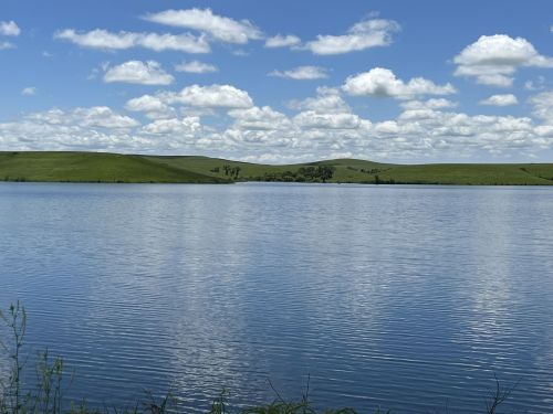 A serene lake surrounded by lush green hills under a bright blue sky with fluffy white clouds.
