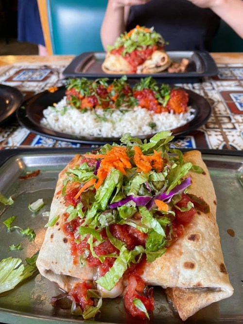 A close-up of a burrito topped with lettuce, salsa, and vegetables, with rice and another dish in the background.