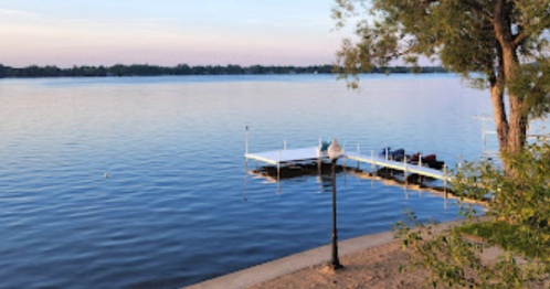 A calm lake at sunset with a wooden dock and a tree in the foreground, reflecting soft colors on the water.