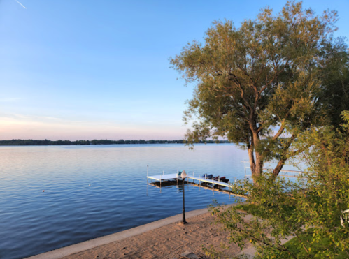 A serene lakeside view with a dock, trees, and calm water under a clear blue sky.