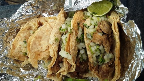 A close-up of four tacos filled with meat, onions, and cilantro, served on foil with a lime wedge.